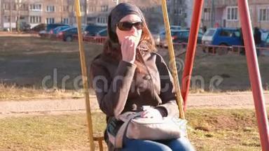 年轻的黑发女人带着太阳镜在秋千上摇摆，在<strong>一座</strong>城市房子的院子里的游乐场里吃华夫饼。