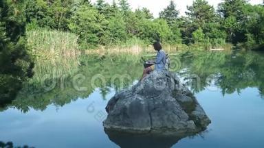 一个年轻的自由职业者坐在湖中央的石头上，手提电脑。 自由职业概念。 出差工作。 无线无线