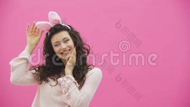 复活节的节日。复活节兔子耳朵上带着微笑的年轻女子在粉色背景上跳来跳去看着复制品