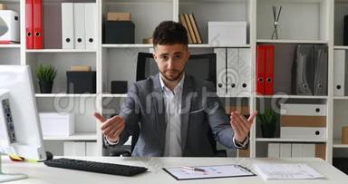 穿着灰色夹克的商人坐在白色办公室的桌子上看着摄像机