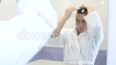 一个年轻人把摩丝涂在头发上。 浴室镜子前穿着白色浴袍的男人.. 查看