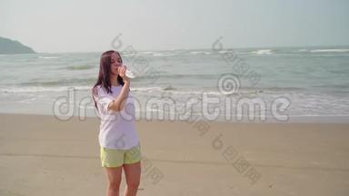 年轻女子从海边的瓶子里喝冷水。 在沙滩上享受清凉矿泉水的成年女士。